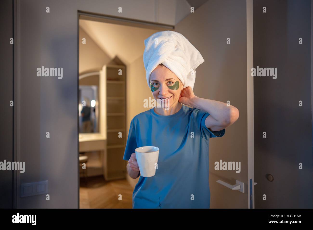 Ruhige Frau mittleren Alters mit Handtuch auf dem Kopf und Flecken unter den Augen, die eine Tasse Kräutertee halten und zu Hause in die Kamera schauen. Selbstpflege am Morgen oder Abend Stockfoto