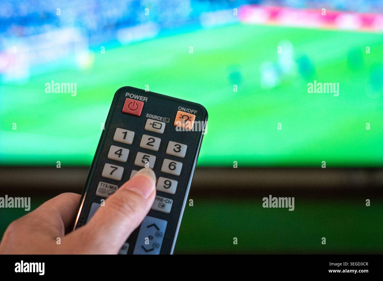 Fußballspiele zu Hause ansehen, Fernbedienung aus nächster Nähe halten, mit unscharfem tv-Bildschirm des Fußballspiels im Hintergrund. Stockfoto