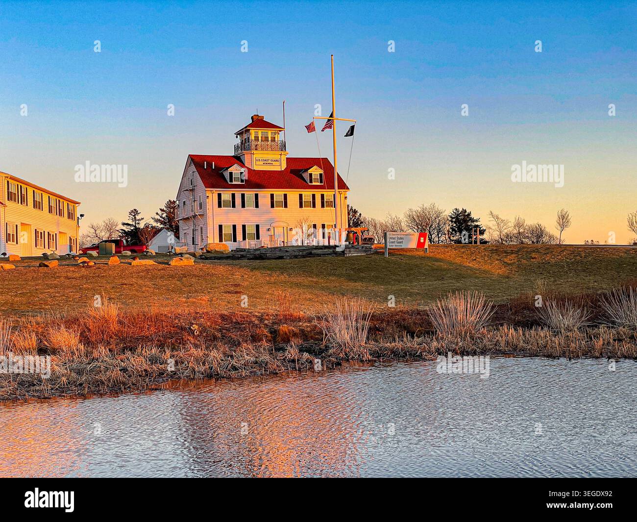Blick auf die US Coast Guard Station Martha's Vineyard mit seinem charakteristischen roten Dach und der weißen Fassade während der goldenen Stunde über einen ruhigen Teich in Edgartown Stockfoto