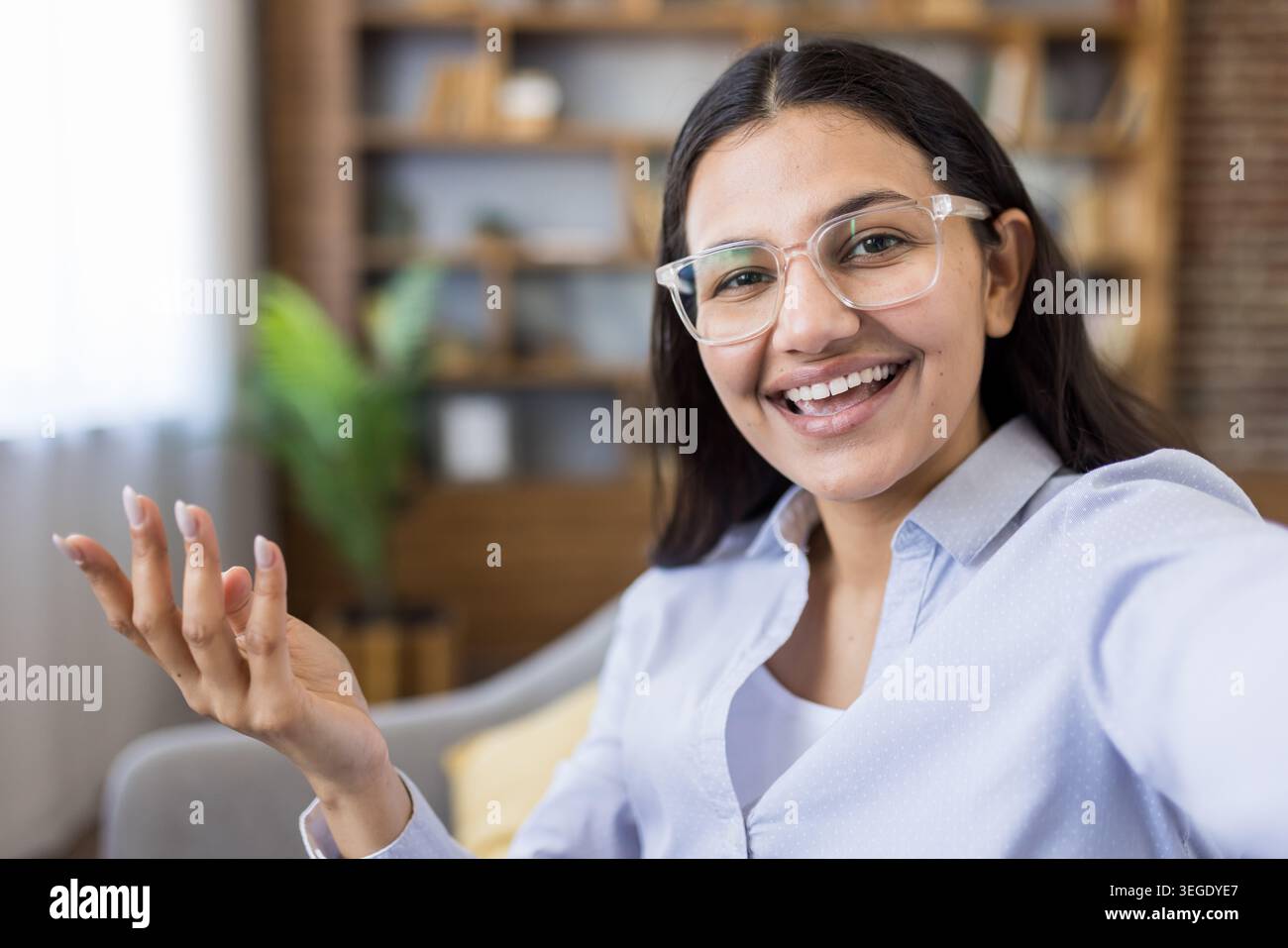 Junge Frau mit Brille kommuniziert gerne aus der Ferne oder fängt glückliche Momente ein, streckt die Hand, während sie direkt in die Kamera blickt, sitzt zuhause Stockfoto