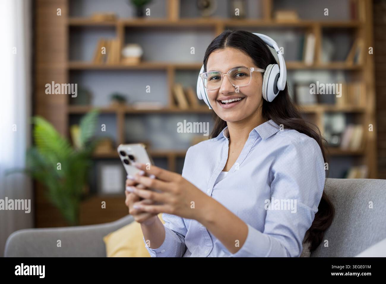 Glückliche junge Frau mit Kopfhörern und Brillen, lächelnd in die Kamera, während sie Musik hört oder eine Online-Lektion mit einem Smartphone im Heimbüro Stockfoto