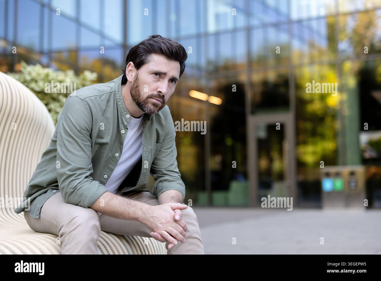 Nachdenklicher Mann mit Bart, der auf einer modernen Bank in einer urbanen Umgebung sitzt und besorgt aussieht, während er vor einem verschwommenen modernen Gebäude gefaltet die Hände legt Stockfoto