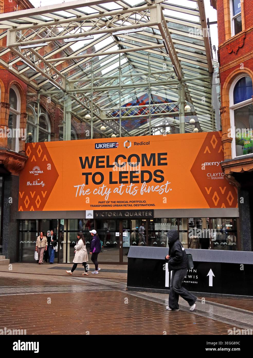 Eingang zum Victoria Quarter Leeds mit orangefarbenem Welcome to Leeds-Schild, Shoppers und Glasarkaden-Baldachin Stockfoto