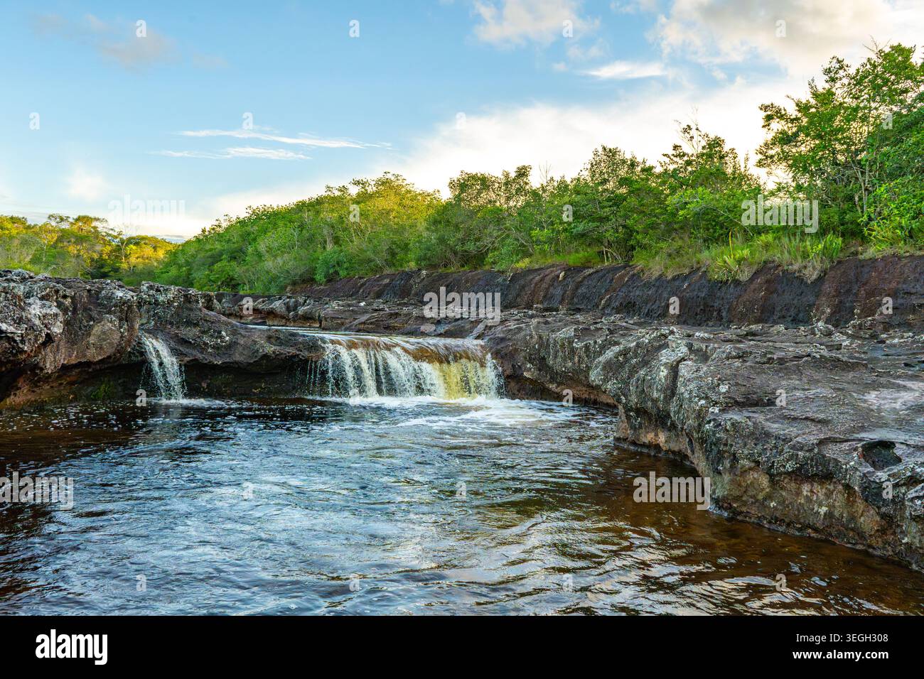 Eine kaskadierende Wasserquelle fließt über felsiges Gelände, umgeben von lebendigen grünen Pflanzen unter einem klaren blauen Himmel in Guaviare. Stockfoto