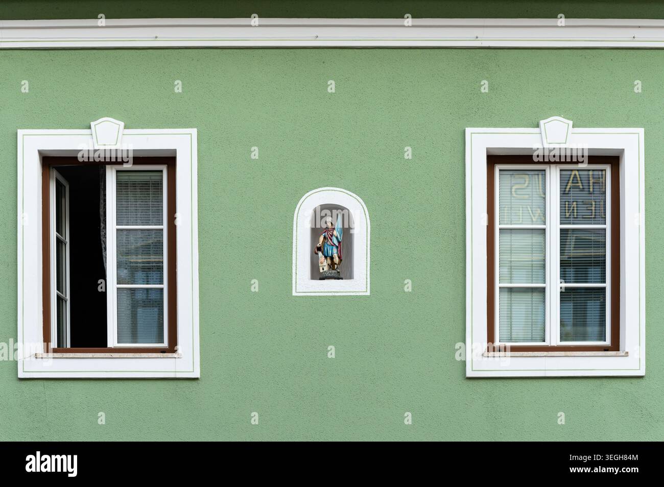 Mariazell, Österreich - 12. Juni 2020: Kleine gemalte heiligenfigur-Schrein-Nische zwischen weiß gerahmten Fenstern auf verwitterter grüner Stuckfassade mit Reflexion Stockfoto