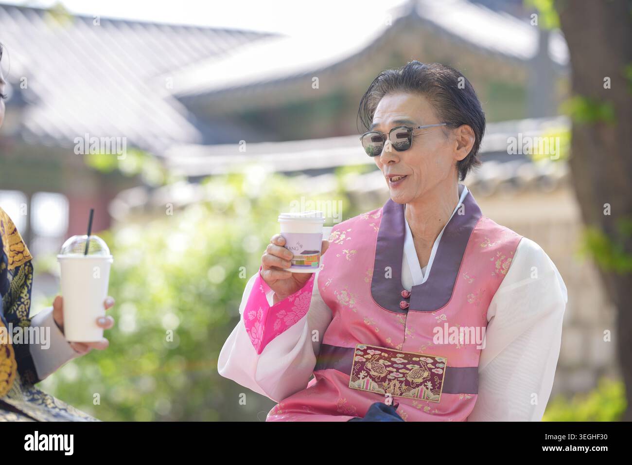 Eine Frau in traditioneller koreanischer Kleidung hält eine Kaffeetasse. Sie trägt eine Sonnenbrille, lächelt sanft. Sanftes Sonnenlicht leuchtet durch Bäume hinter ihr. Hintergrundinformationen Stockfoto