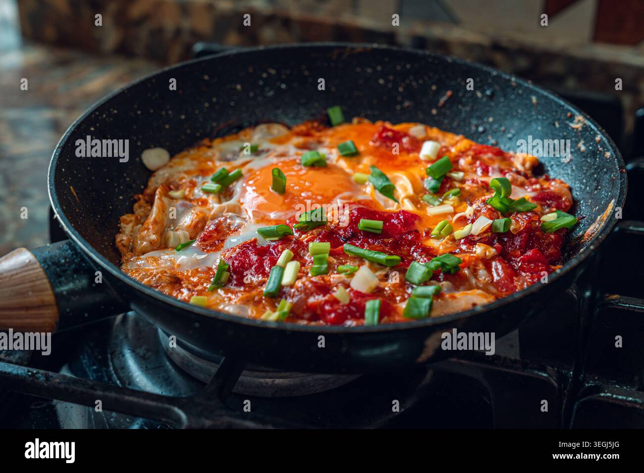 Eine Nahaufnahme eines lebhaften Eiergerichts, das in einer Pfanne zubereitet wird, garniert mit frischen grünen Zwiebeln und Tomaten, wodurch eine farbenfrohe und appetitliche Mahlzeit entsteht. Stockfoto