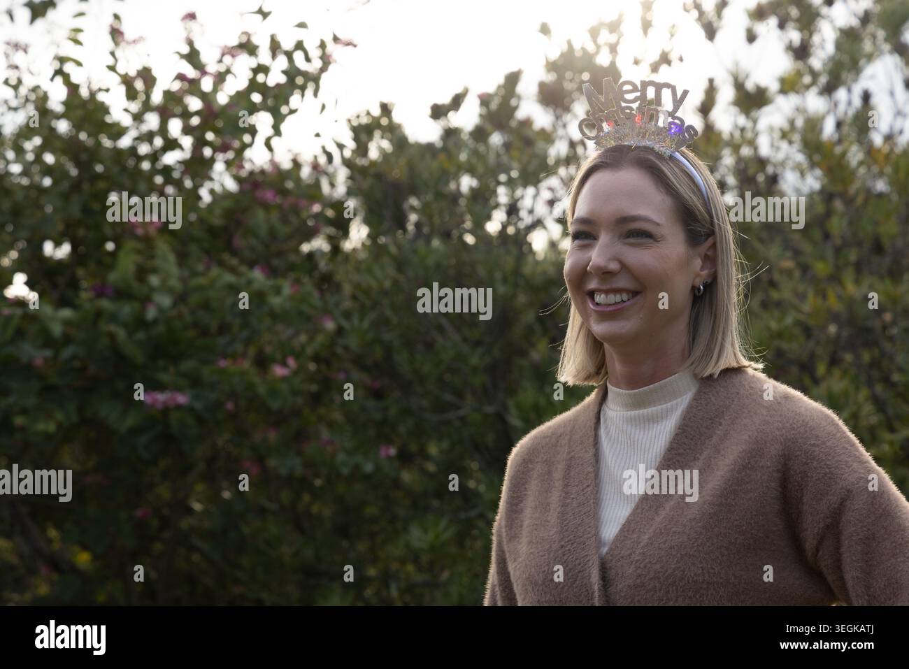 Frau lächelt, trägt glitzerndes, fröhliches Stirnband und Strickjacke im Garten mit rosa Blumen, Kopierraum Stockfoto