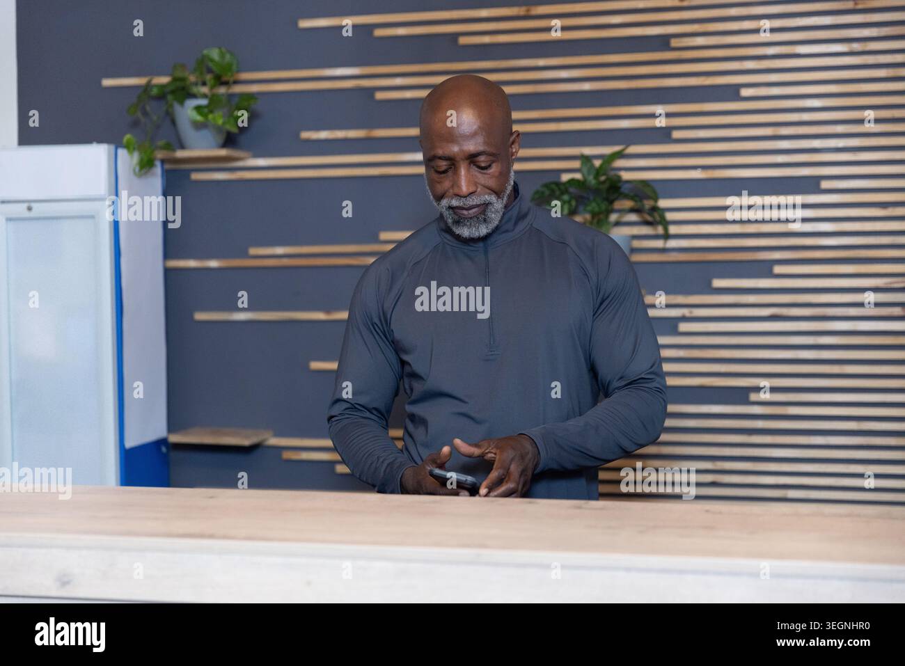 Senior Afroamerikaner, der das Smartphone überprüft, steht hinter Holztheke in einem dunklen Pullover mit Reißverschluss Stockfoto