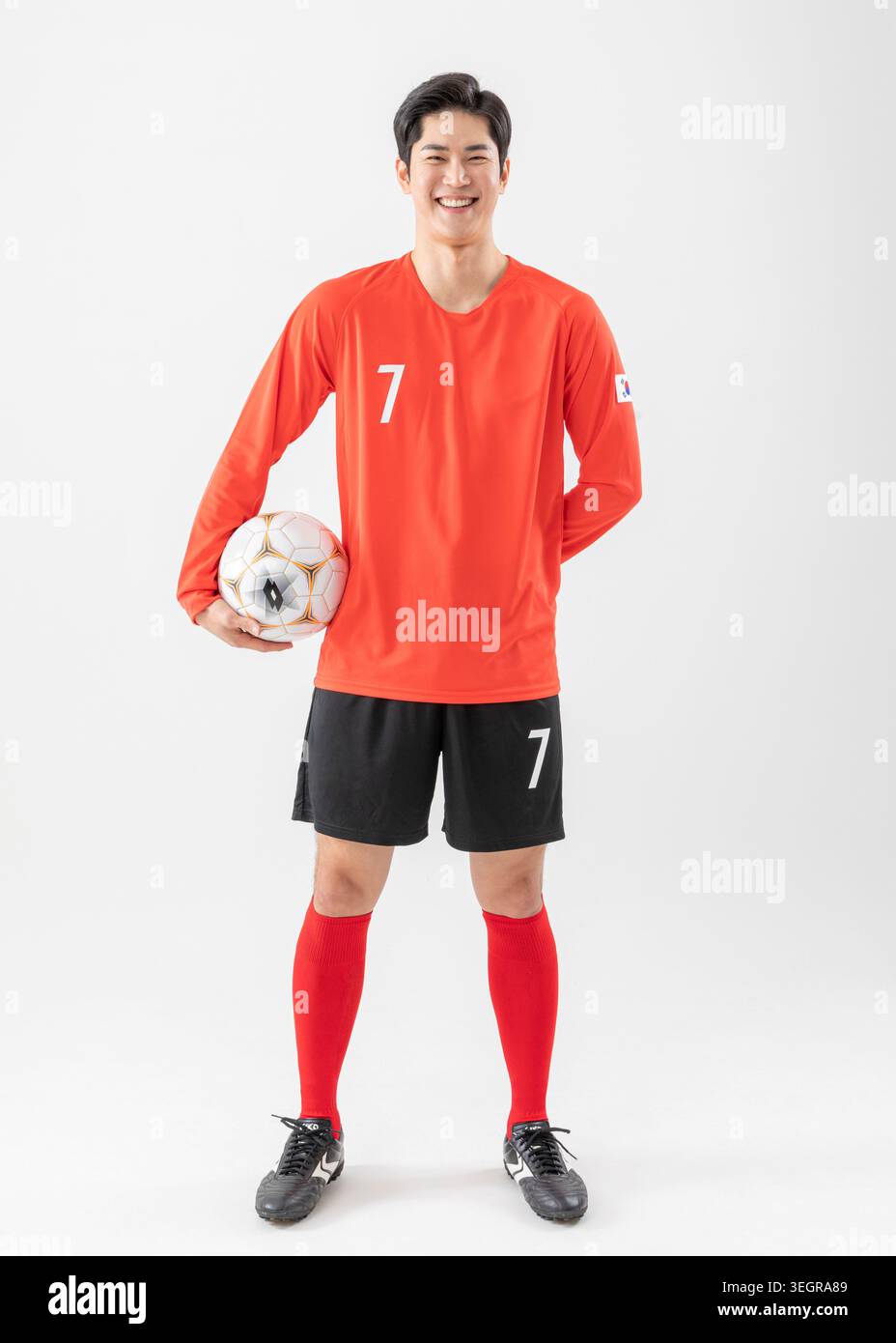 Ein junger männlicher Fußballspieler steht selbstbewusst und hält einen Fußball vor weißem Hintergrund. Stockfoto