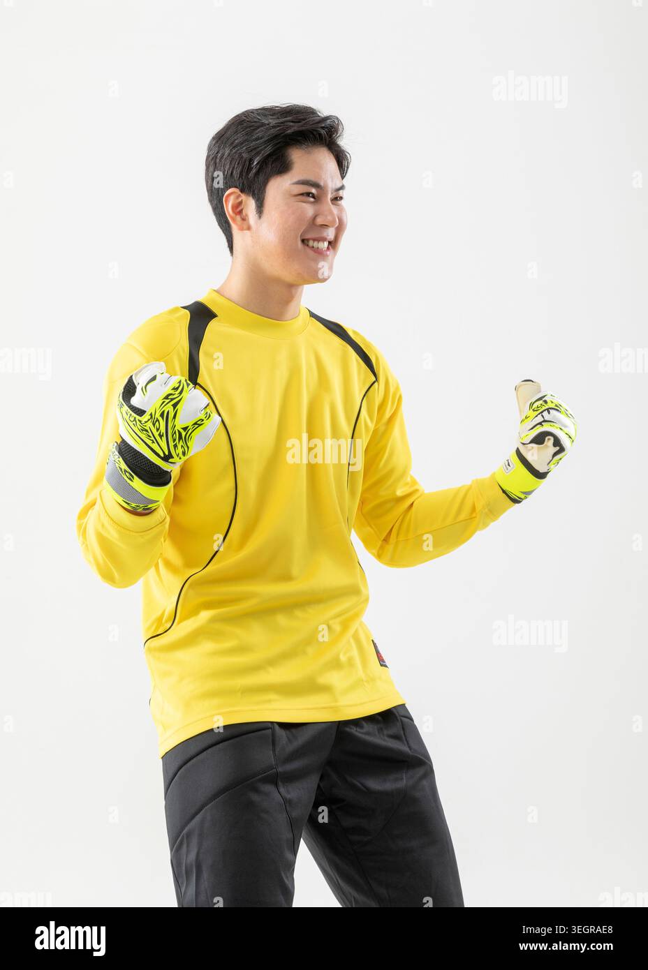 Ein junger asiatischer Torhüter steht mit triumphalen Armen in einem gelben Trikot und passenden Handschuhen vor weißem Hintergrund. Stockfoto
