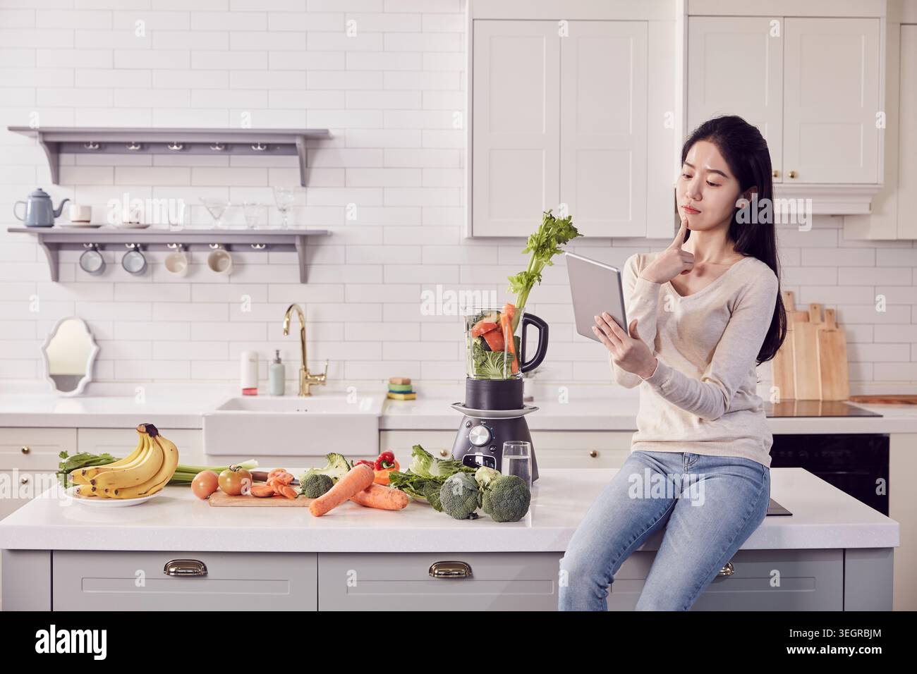 Eine Frau sitzt auf einer Kücheninsel und sucht mit einem Tablet nach Rezepten, umgeben von Zutaten und einem Mixer. Stockfoto