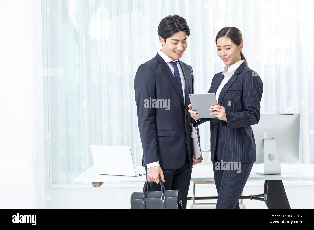 Eine junge asiatische Geschäftsfrau und ein junger asiatischer Geschäftsmann in einem modernen Büro, beide lächeln, während sie gemeinsam ein Tablet überprüfen. Stockfoto