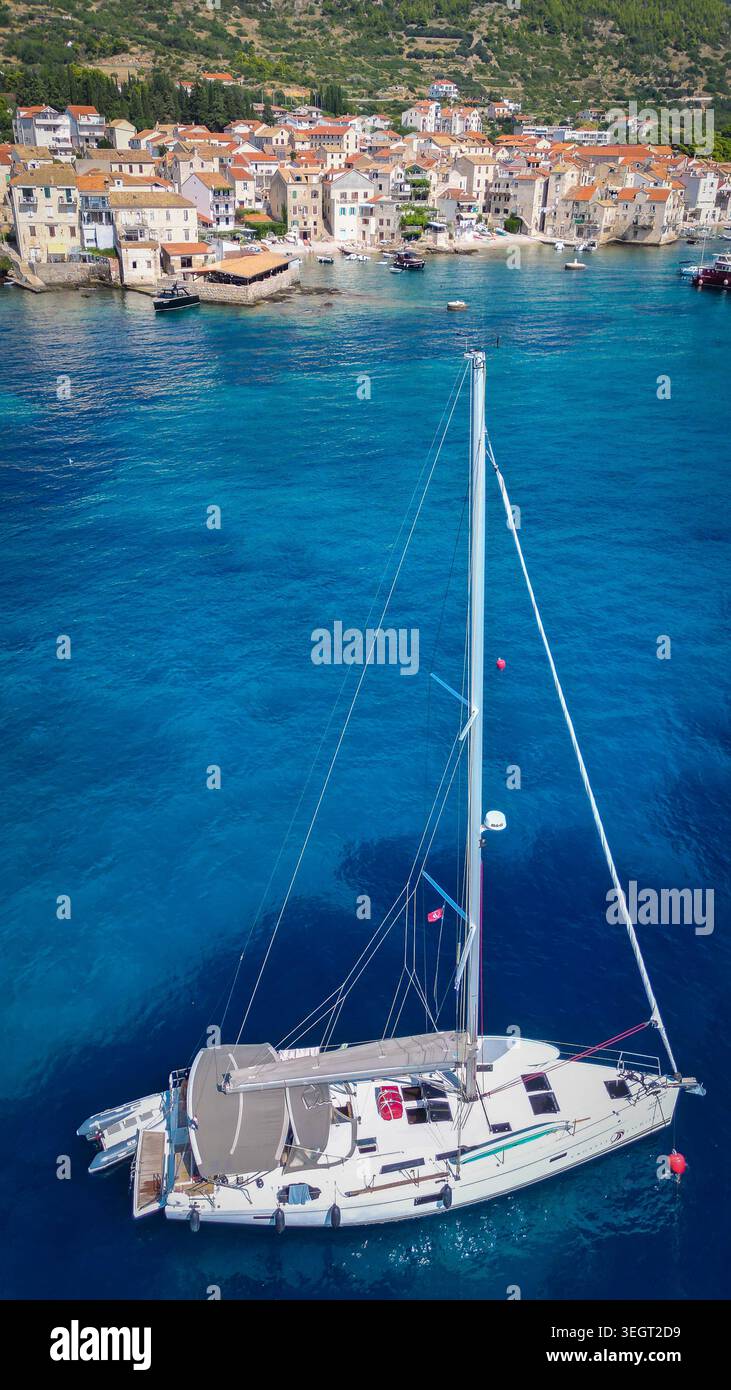 Aus der Vogelperspektive auf ein weißes Segelboot, das im türkisfarbenen Wasser ankert, mit der historischen Küstenstadt und den üppigen grünen Hügeln in Komiza, County Split-Dalmatia, Cro Stockfoto