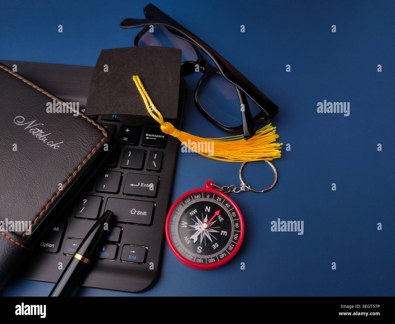 Eine Sammlung von Gegenständen, einschließlich Notizbuch, Stift, Tastatur, Graduierungskappe, Brille, und ein Kompasslayout auf Blau. Stockfoto