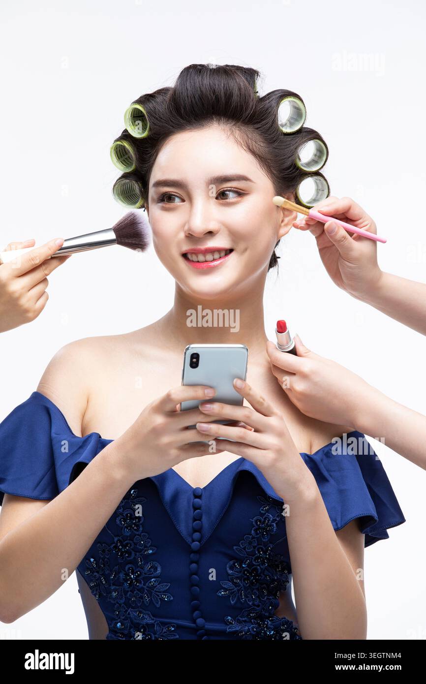 Eine Frau ist von mehreren Händen umgeben, die Kosmetikwerkzeuge und -Produkte halten, während sie ein Telefon hält und lächelt. Stockfoto