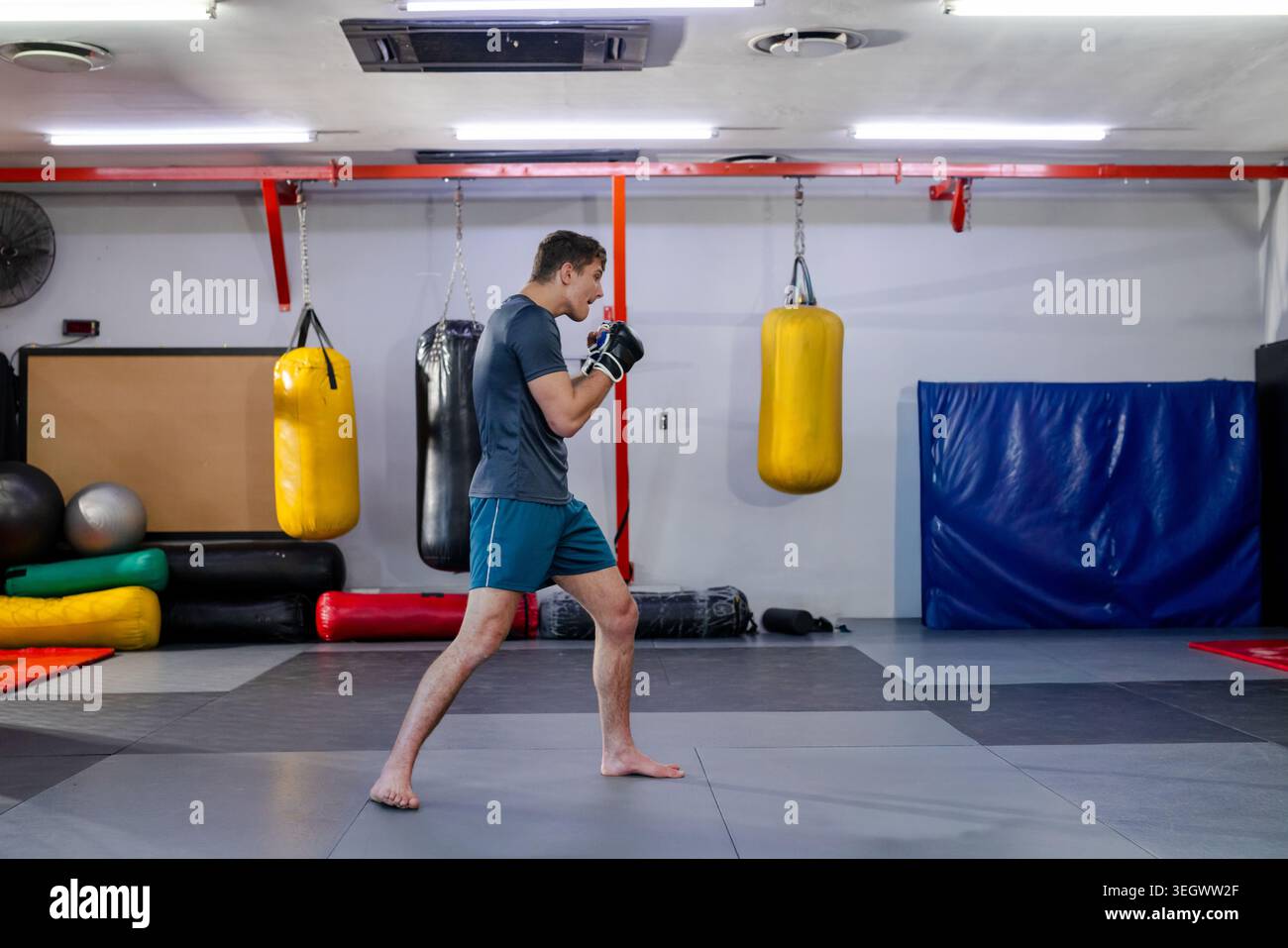 Mann, der Boxen mit schwarzen Handschuhen übt, Wache hält und in der Nähe gelber schwerer Taschen ins Fitnessstudio tritt Stockfoto