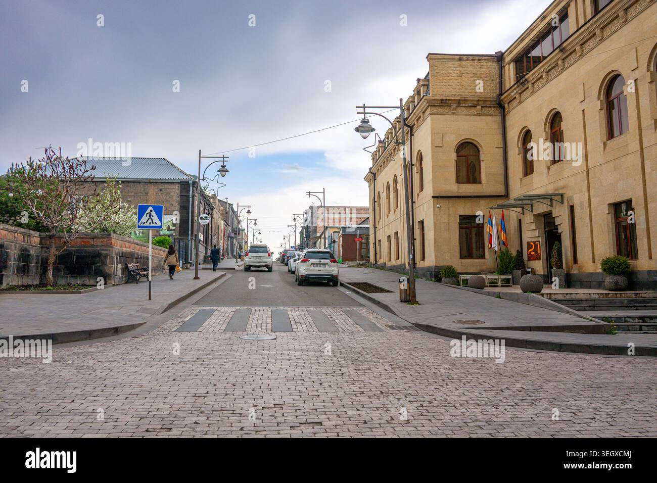 Die kopfsteingepflasterte Straße erstreckt sich zwischen historischen Gebäuden unter bewölktem Himmel. Die Fahrzeuge fahren langsam auf einer ruhigen Stadtstraße Stockfoto