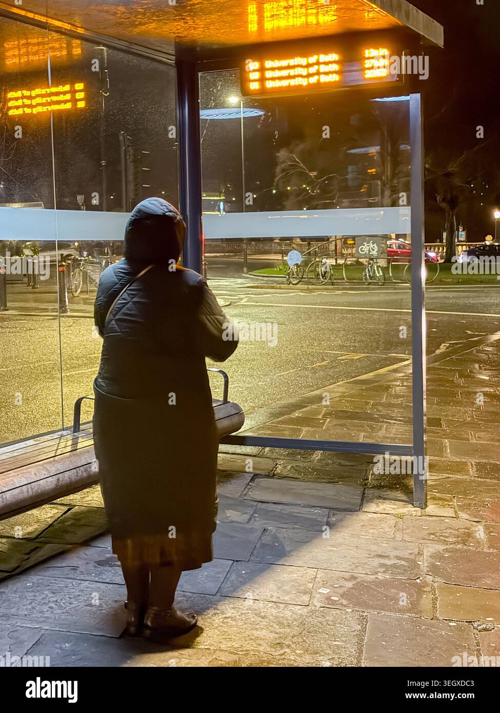 Bath, Somerset, England, Großbritannien - 17. Dezember 2025: Frau allein in der Nacht, die an einer Bushaltestelle auf einen Bus wartet. - Smartphone-aufgenommenes Stockfoto