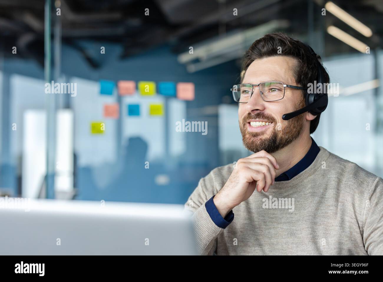 Glücklicher bärtiger Mann mit Brille und Headset, der am Laptop arbeitet, Unterstützung und Unterstützung bietet und ein positives Kundenerlebnis in einer modernen Büroumgebung verkörpert Stockfoto