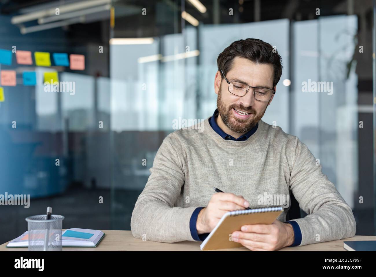 Junger, lächelnder Geschäftsmann mit Brille, der an einem modernen Schreibtisch in ein Spiralnotizbuch schreibt und sich auf Brainstorming, die Planung von Aufgaben und die Organisation von Ideen für ein Projekt oder eine Besprechung konzentriert Stockfoto