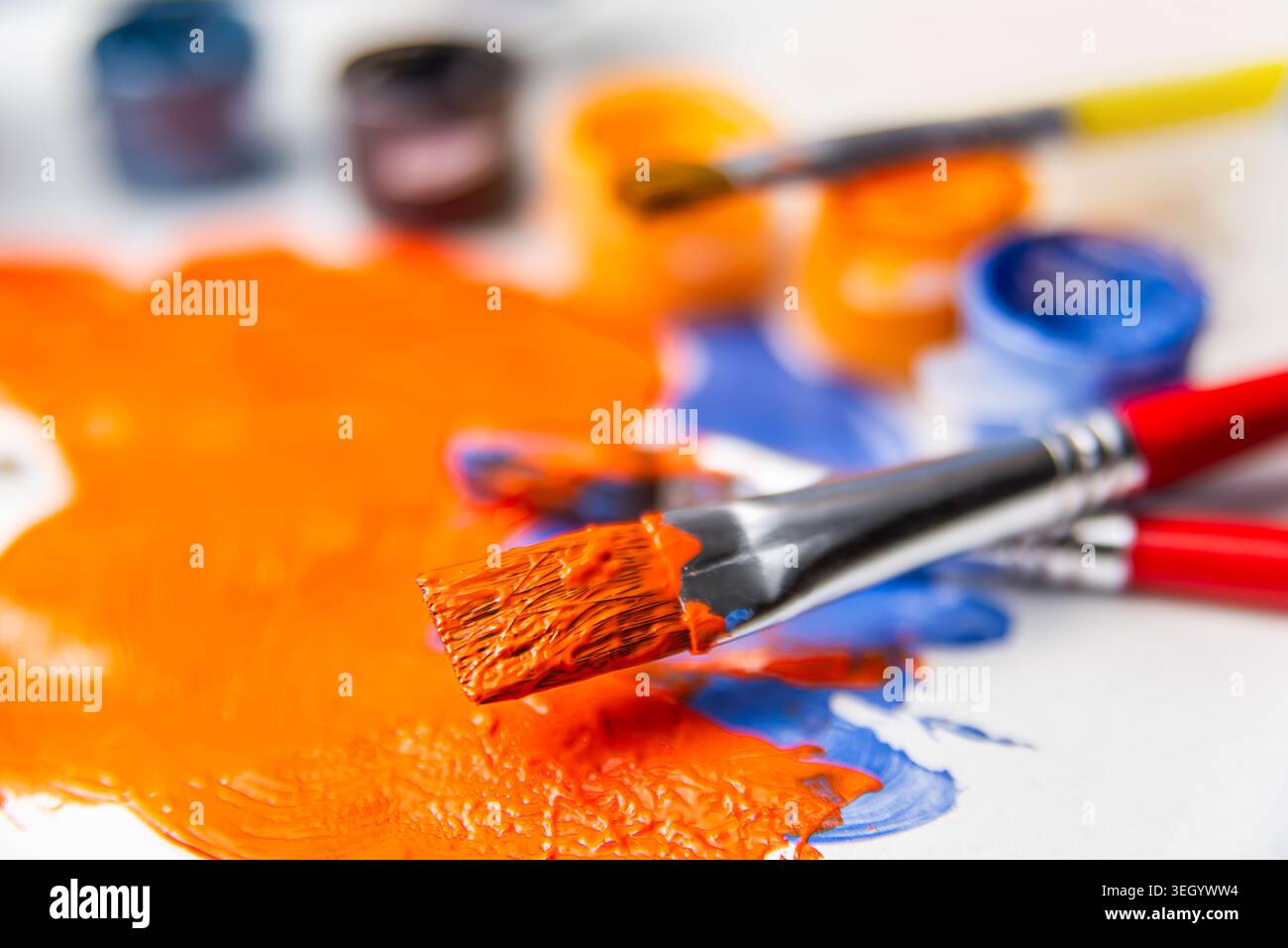 Hellorange und blaue Farbe bedeckt eine Palette mit Pinseln in der Nähe. Künstler kreieren an einem Tisch, der mit Farbzubehör gefüllt ist. Stockfoto