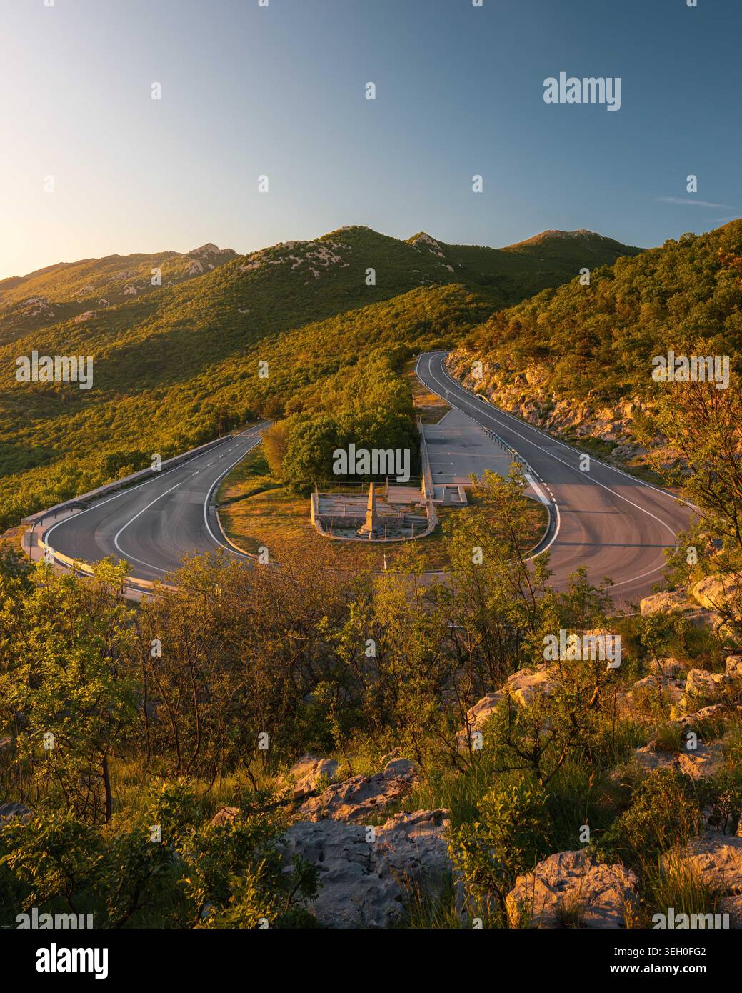 Aus der Vogelperspektive auf eine gewundene Bergstraße mit einer scharfen Haarnadelkurve und einem Steindenkmal umgeben von üppigen grünen Hügeln während der goldenen Stunde in Rijeka, Pri Stockfoto