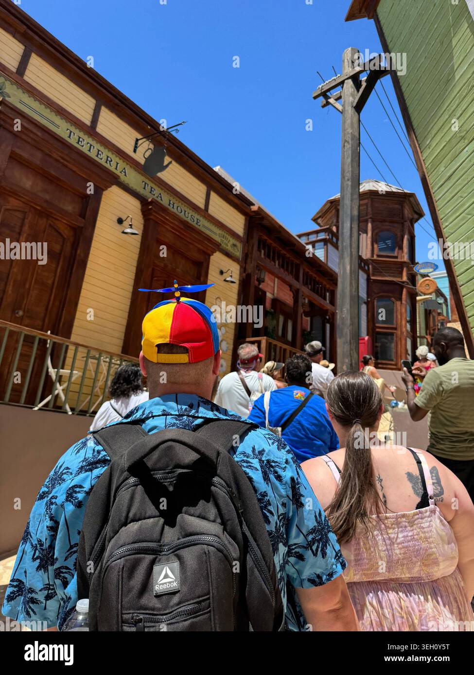 Gruppe von Touristen, die durch eine Outdoor-Freizeitpark-Gasse laufen oder in der Schlange neben rustikalen Gebäuden mit einem Mann in einem lustigen Hubschrauberhut stehen - Smartphone-aufgenommenes Stockfoto