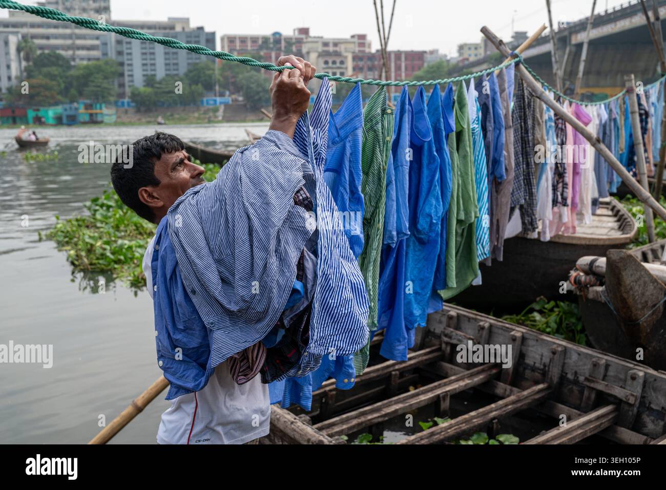 Dhaka Division, Bangladesch - 14. Mai 2026: Blick auf einen Mann, der seine Wäsche an einem Seil über einem Holzboot auf dem Buriganga River mit städtischen Gebäuden in Th hängt Stockfoto