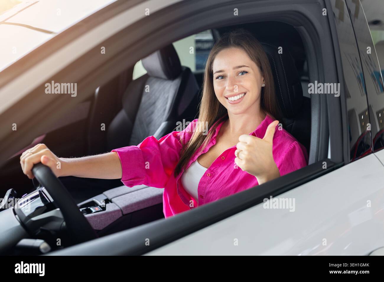 Glückliche junge Frau, die im Auto sitzt und im Autohändler die Daumen hochlegt Stockfoto