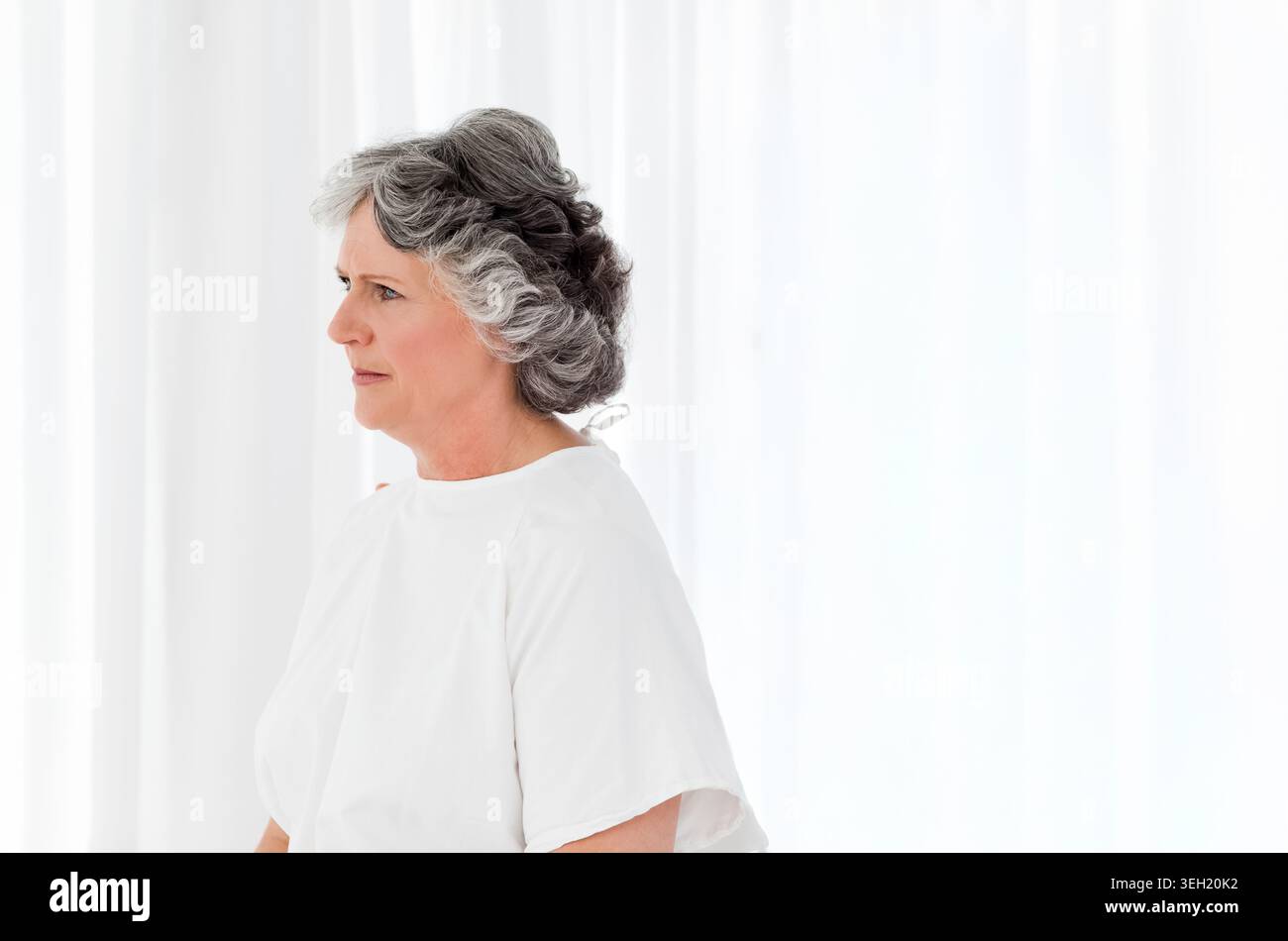 Eine ältere Frau, die im Profil steht und ein weißes Kleid trägt, sieht von transparenten Vorhängen links, Kopierraum Stockfoto
