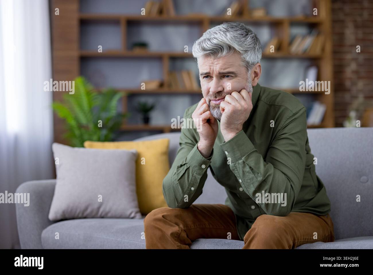Reifer Mann mit grauen Haaren, der nachdenklichen Ausdruck und Traurigkeit zeigt, allein auf einem Sofa zu Hause sitzt und Gefühle von Depression, Angst und Einsamkeit erlebt Stockfoto