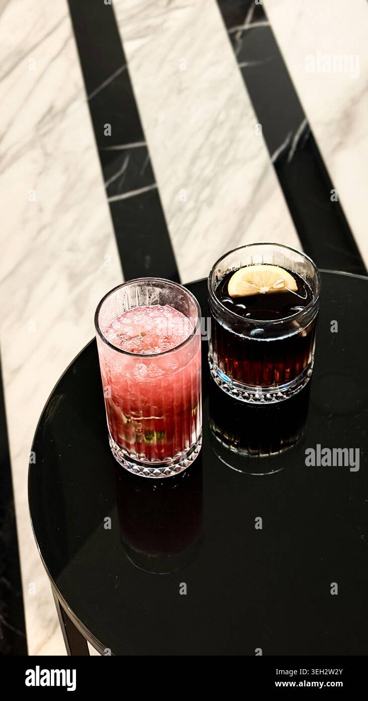 Zwei Cocktailgläser auf einem schwarzen Marmortisch, eines mit rosafarbenem Getränk mit Minze und das andere mit dunklem Getränk und Zitronenscheibe Stockfoto