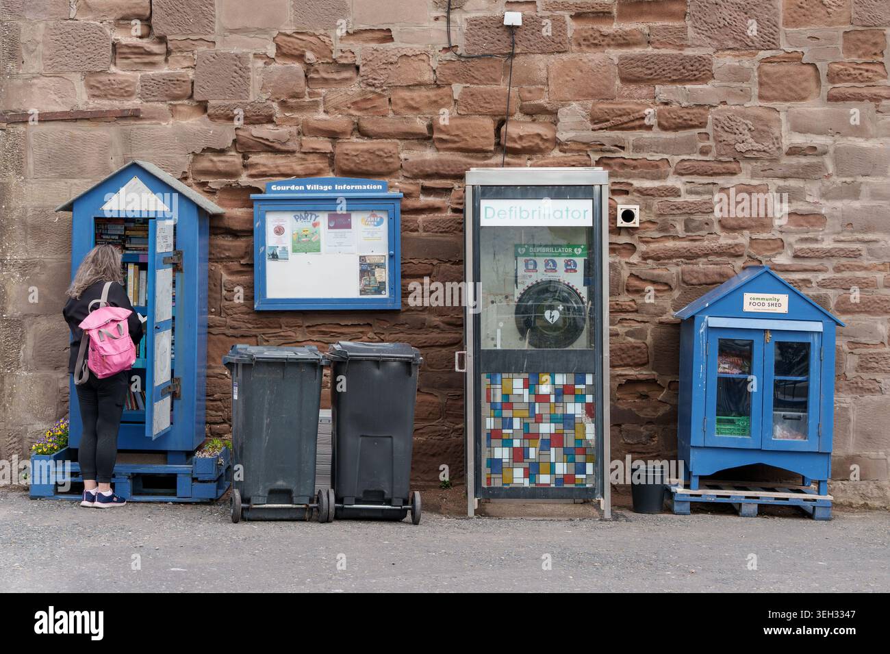 Frau durchsucht Bücher im Gourdon-Buchstand. Neben ihr steht ein Gemeinschaftsrestaurant, ein öffentlicher Defibrillator und eine Dorfinformationstafel, A Stockfoto