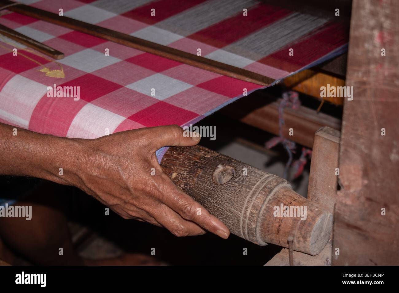 Nahaufnahme eines Handwerkers, der traditionellen Jamdani-Stoff in Rot-weiß-Karomuster auf einem hölzernen Handwebstuhl webt und handgefertigte Textilien zeigt. Stockfoto