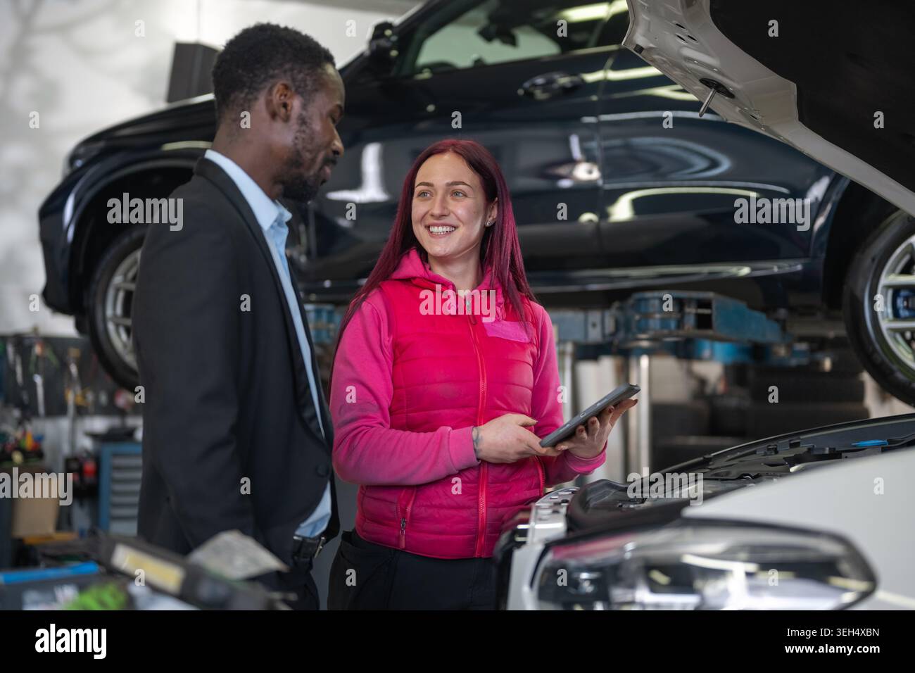 Eine Mechanikerin erklärt einem Kunden, einem afroamerikanischen Geschäftsmann, eine Checkliste für die Fahrzeugwartung auf dem Tablet-Bildschirm. Stockfoto