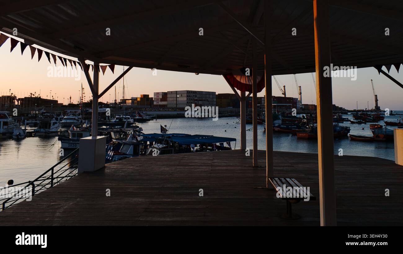 Malerischer Blick auf einen belebten Hafen mit Fischerbooten und Frachtcontainern bei Sonnenuntergang, eingerahmt von der Silhouette eines hölzernen Pavillons am Wasser in Chile. Stockfoto