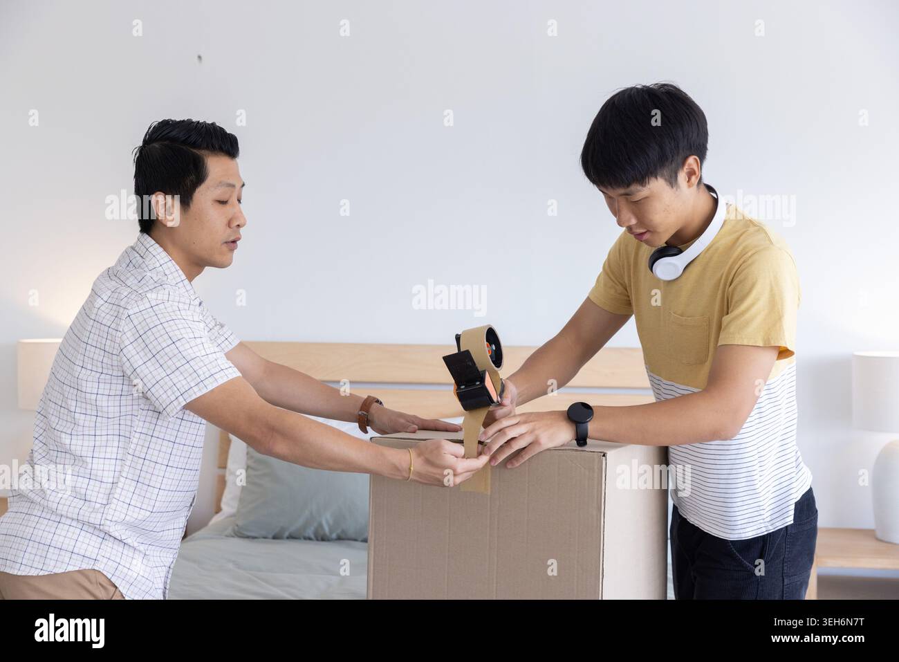 Asiatische Männer, die im Schlafzimmer stehen, versiegeln große Karton-Umzugskarton mit handgehaltenem Bandspender Stockfoto
