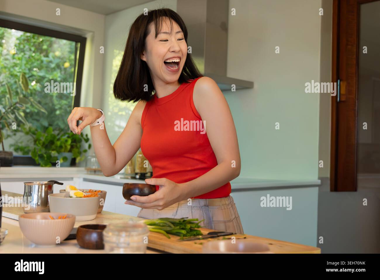 Chinesin trägt rote Spitze, lacht und würzt Essen auf der Kücheninsel, hält eine Holzschale Stockfoto