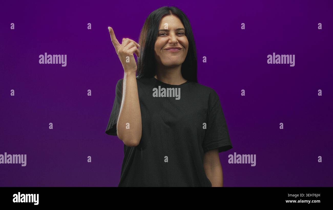 Frau zeigt den Zeigefinger nach oben in Richtung Tempel im Studio mit violettem Hintergrund und dezentem Lächeln; Inspiration für Selbstvertrauen. Stockfoto
