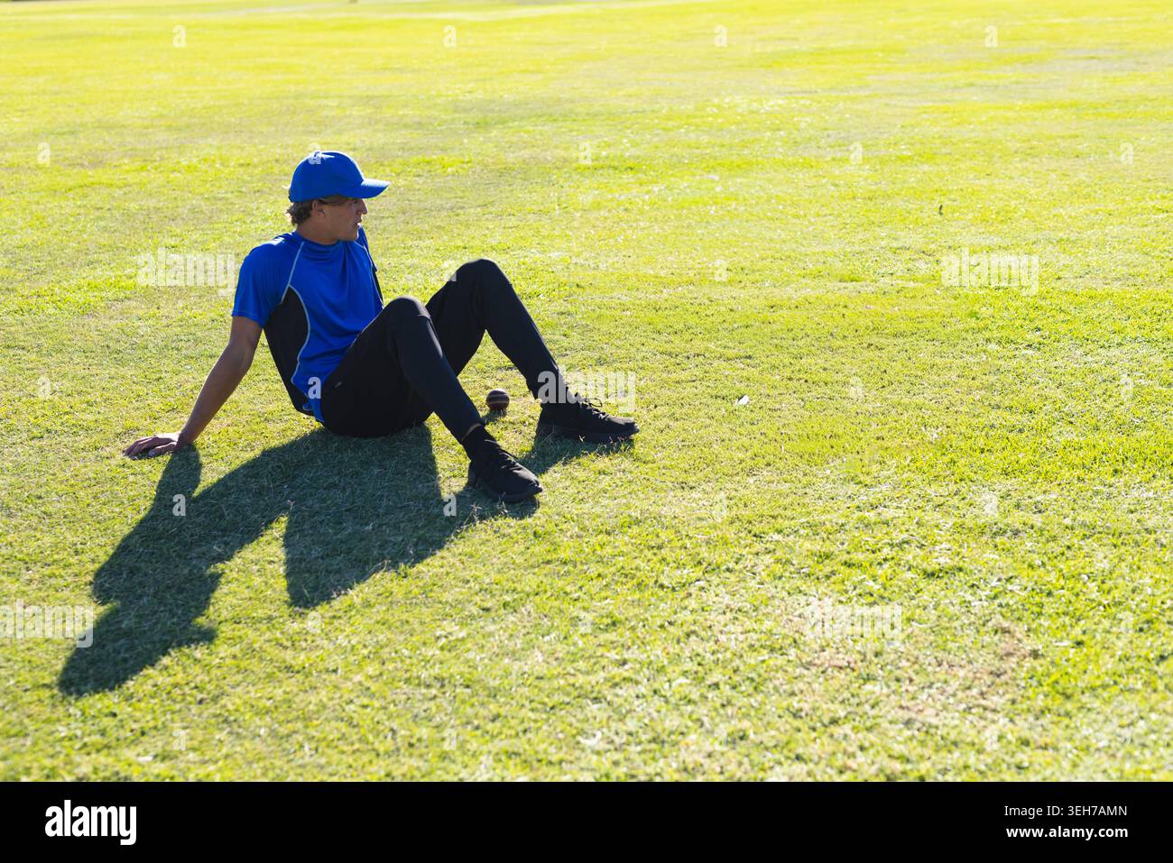 Männlicher Athlet, der auf einem grasbewachsenen Feld sitzt, blaues Hemd und Mütze neben Baseball trägt und Schatten wirft Stockfoto