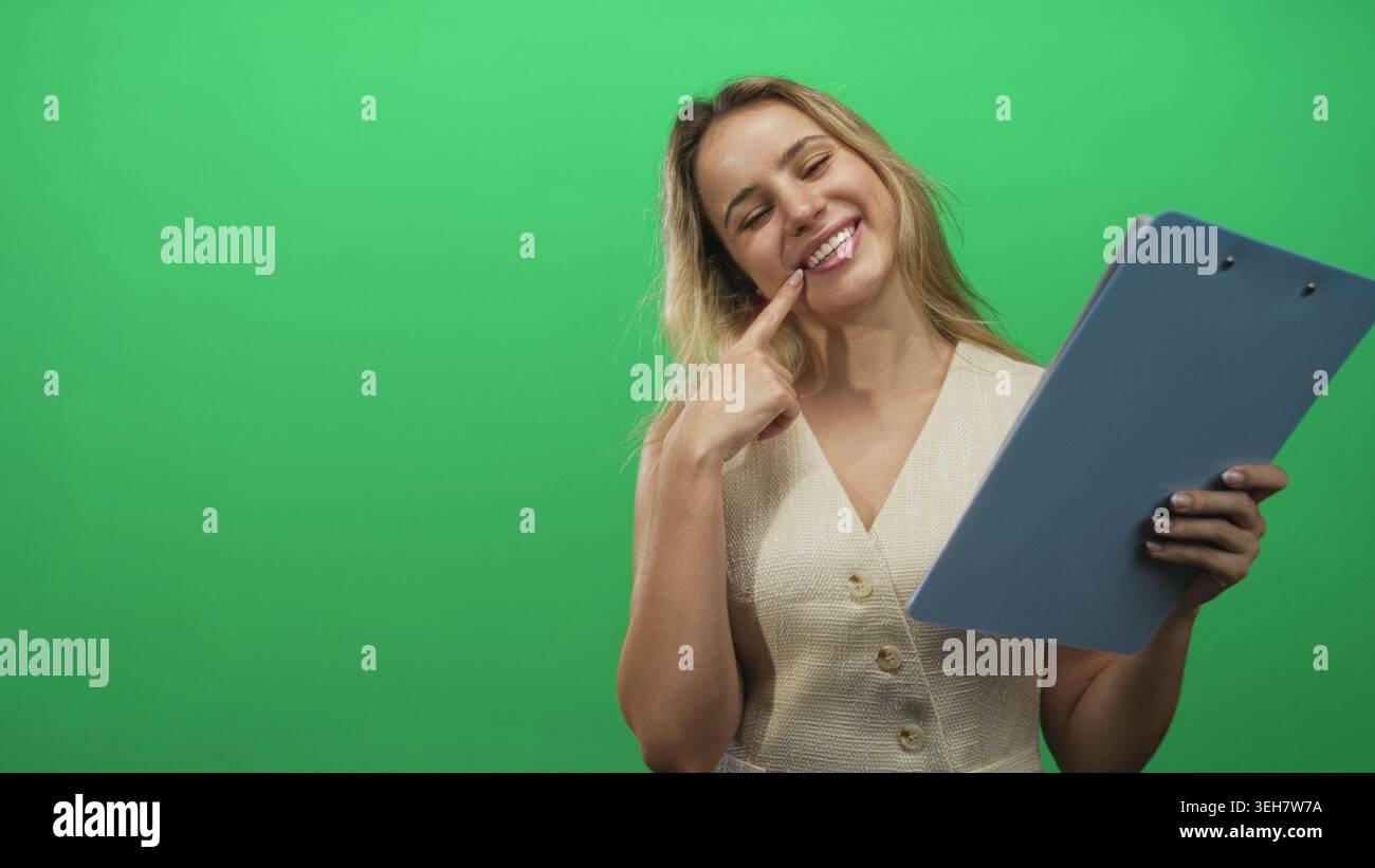 Blonde junge Frau, die das Klemmbrett hält und mit dem Finger nach oben zeigt, während sie im Studio lächelt; verspielte Neugier. Stockfoto
