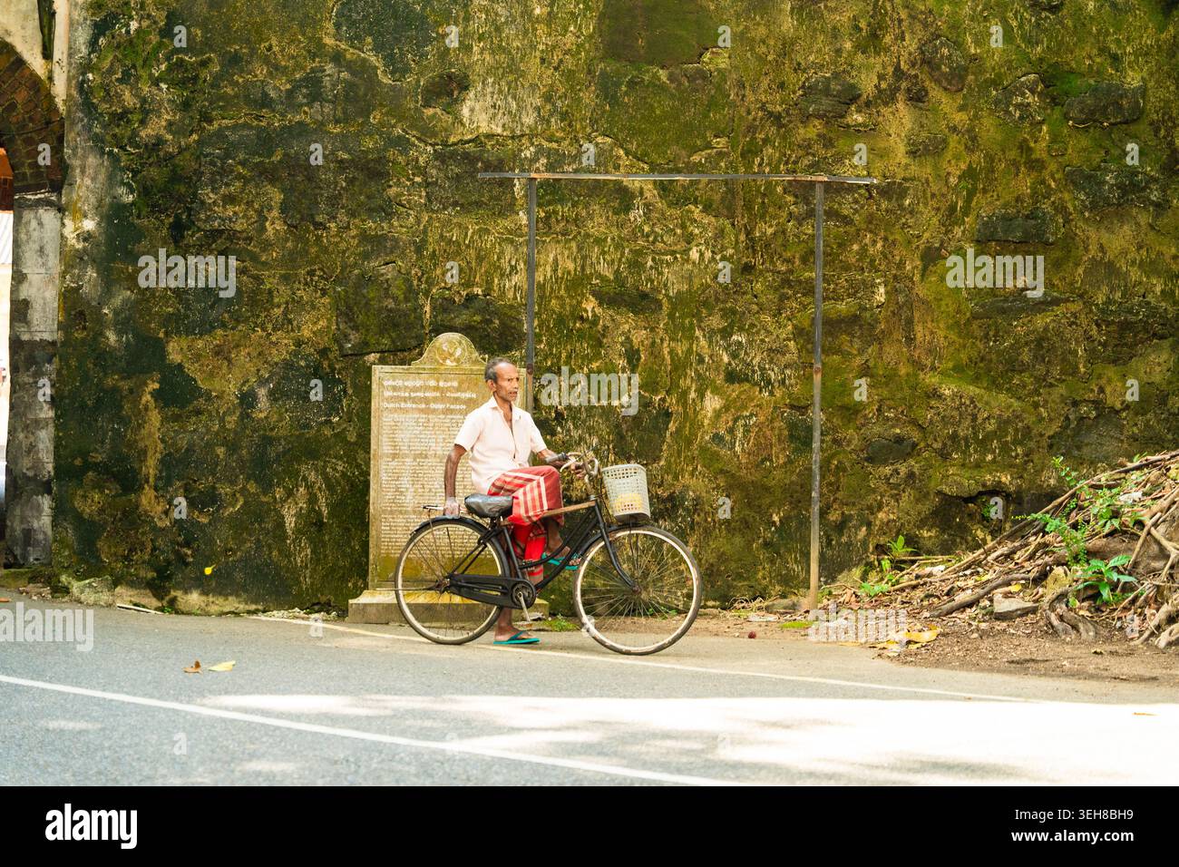 Sri Lanka - 4. Februar 2026: Blick auf einen älteren Mann, der auf einem Fahrrad vor den historischen moosigen Steinmauern des Galle Fort sitzt. Stockfoto