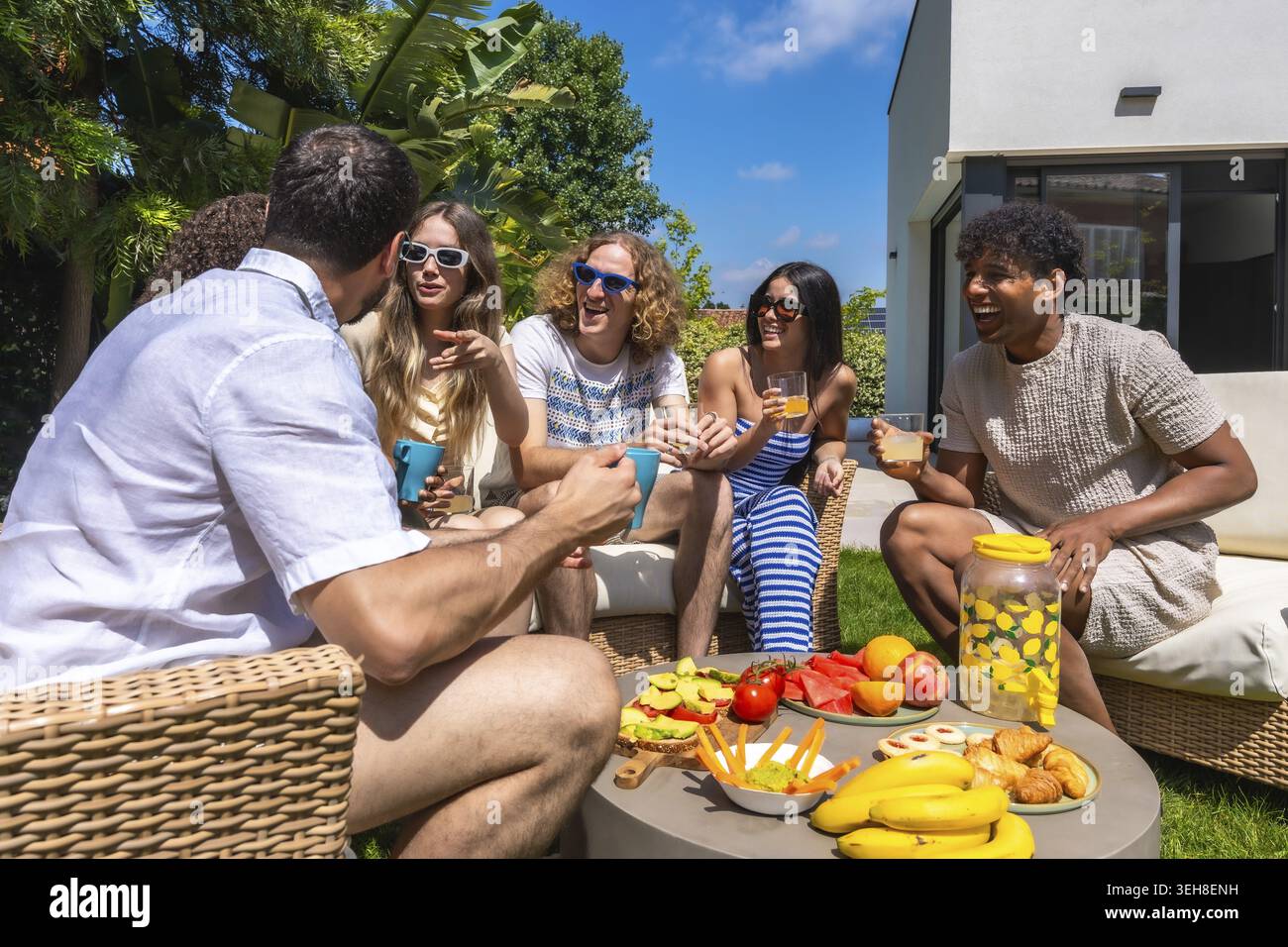 Vielfältige Gruppe junger erwachsener Freunde lachen und unterhalten, während sie sich auf einer Gartenparty im Freien unterhalten, Getränke und gesundes Essen mit modernem A genießen Stockfoto