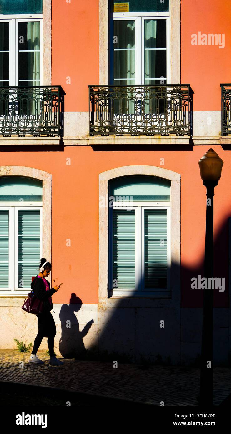 Frau auf Smartphone in dramatischem Schatten vor leuchtender orangener Fassade, Alcântara, Lissabon, Portugal Stockfoto