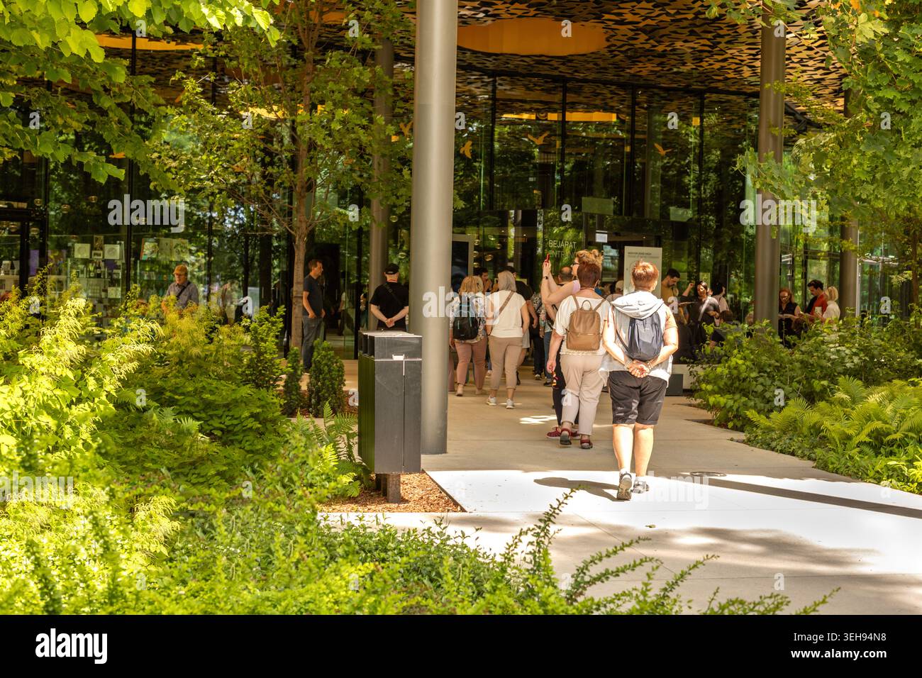 Touristen laufen in der Nähe eines modernen Glasgebäudes in einem üppig grünen Park. Stockfoto