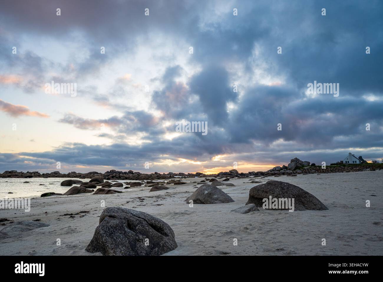 Ein malerischer Sonnenaufgang beleuchtet den Meneham Strand in der Bretagne. Große Granitblöcke sind auf weißem Sand verstreut, während im B ein typisches bretonisches Haus erscheint Stockfoto