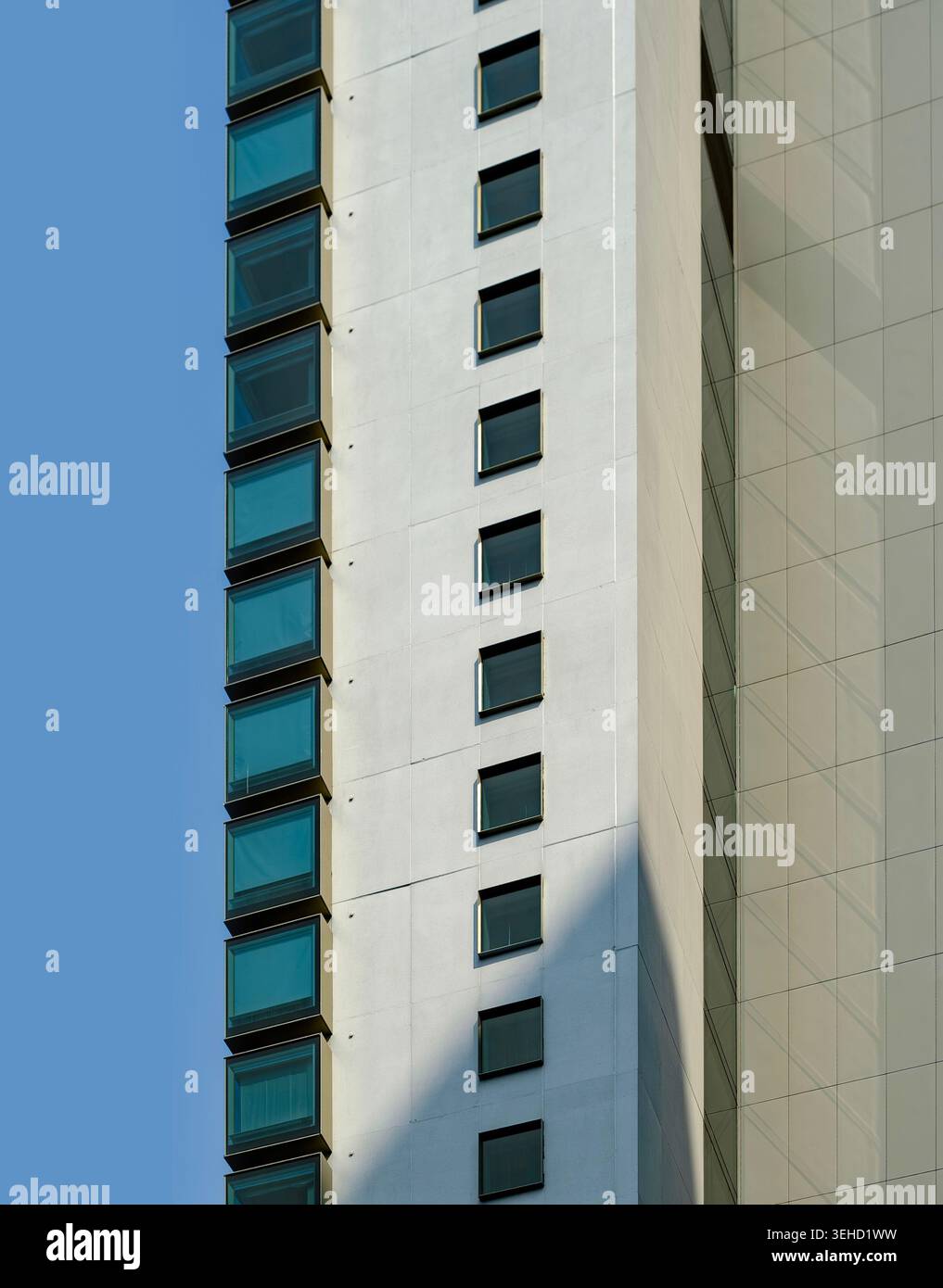 Ein klares, vertikales architektonisches Detail eines weißen Wolkenkratzers mit blau getönten Fenstern, geometrischen Mustern und scharfen Schatten vor einem klaren Himmel Stockfoto