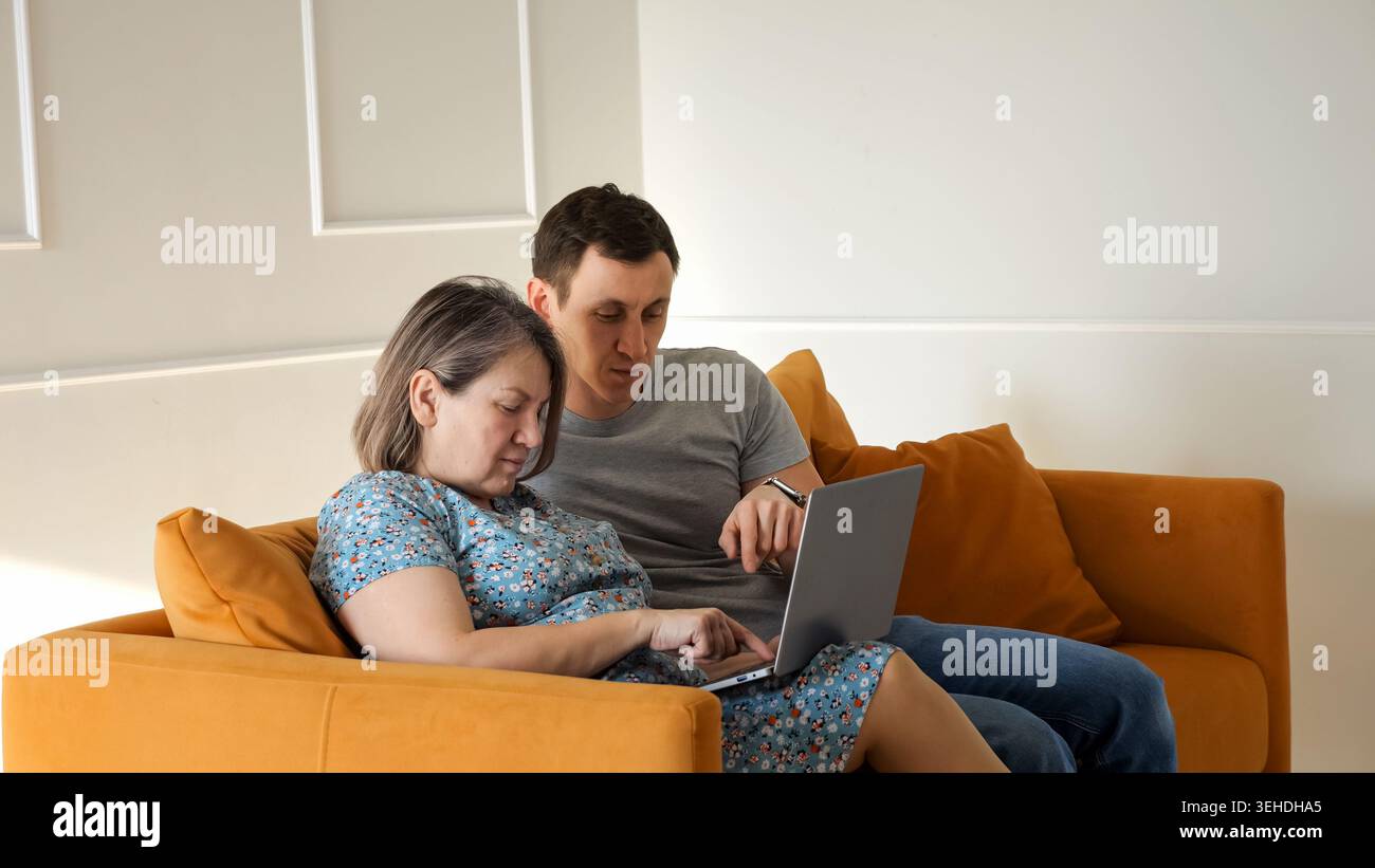 Der Sohn hilft seiner älteren Mutter, neue Technologien und Computerfähigkeiten auf einem Laptop zu erlernen, während er sich auf einem gemütlichen Sofa in einem modernen Wohnzimmer entspannt Stockfoto