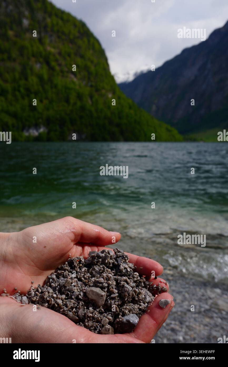 Hände halten nasse Kieselsteine an einem klaren Bergsee, umgeben von steilen grünen Bergen Stockfoto